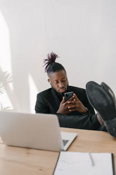 Stylish young man in office using smartphone, feet on desk, relaxed atmosphere.