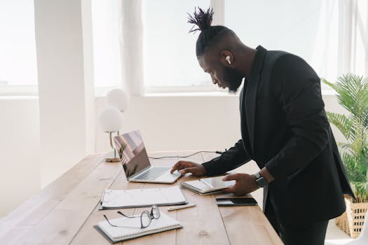 A businessman in a modern office setting, deeply focused on work with his laptop and notepad.