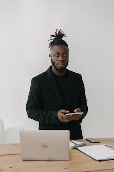 Professional black businessman in business attire standing with a smartphone by a desk in an office.