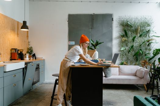 A young woman in casual wear working on a laptop in a modern kitchen with indoor plants.