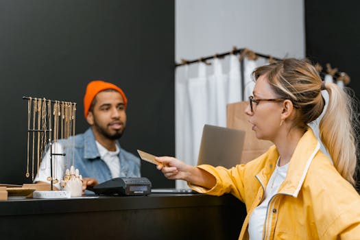 Woman in yellow jacket pays with credit card at shop counter with jewelry display.