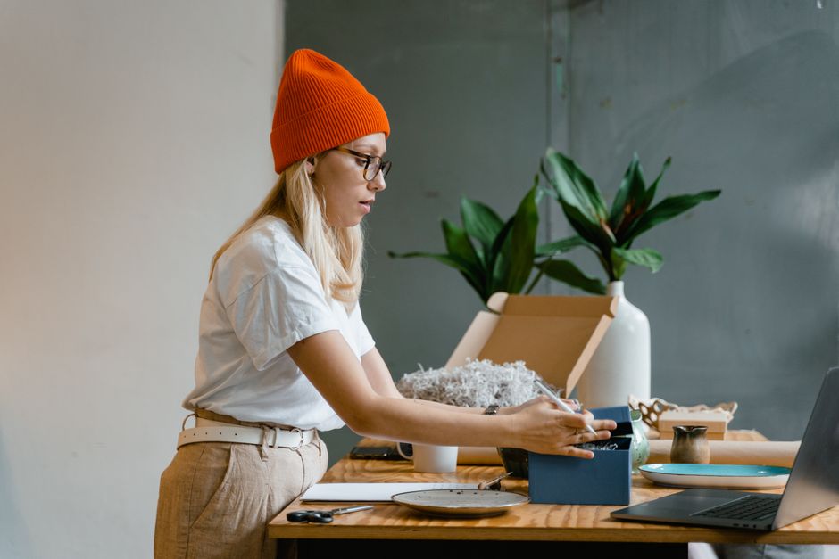 Woman in orange beanie packing gifts at a wooden table with plants in an indoor setting.