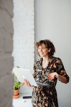 Happy woman in floral dress using a laptop and earbuds indoors, enjoying telecommuting with a bright smile.