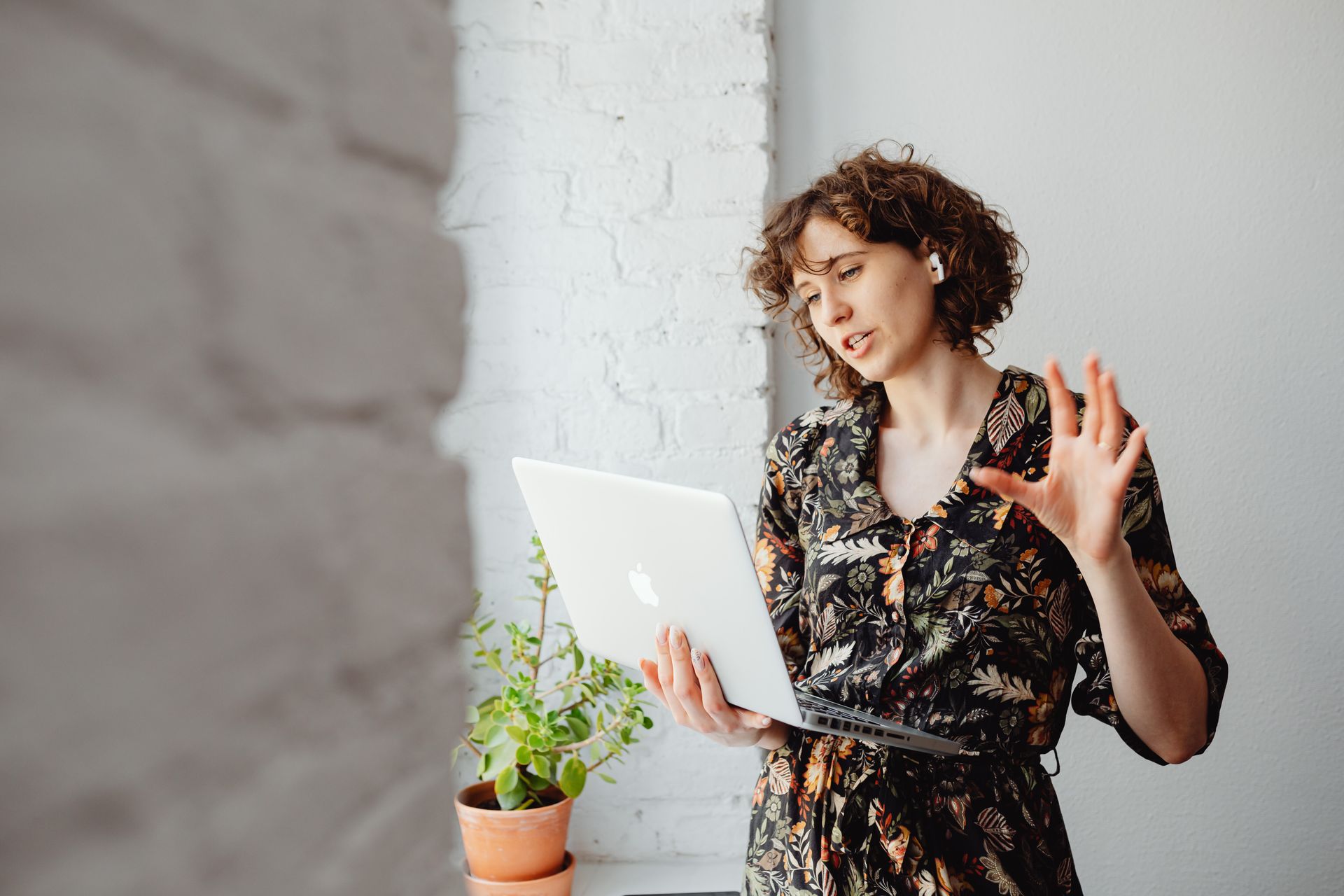 A woman with curly hair conducts an online meeting using her laptop indoors, wearing wireless headphones.