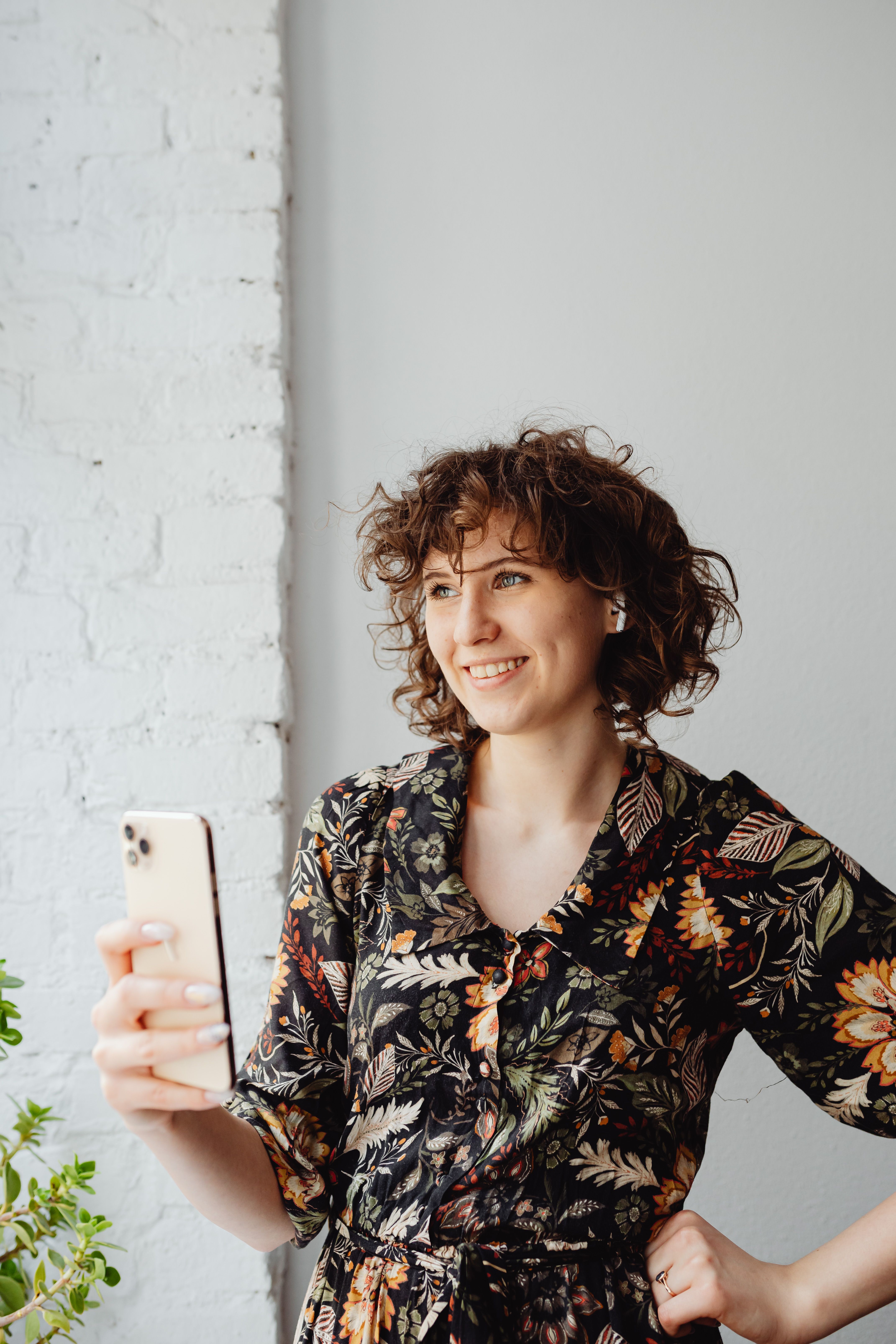 Happy woman with curly hair taking a selfie indoors wearing floral dress and earpods.