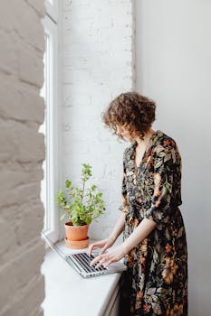 A woman in a floral dress is working on a laptop by a window with a potted plant.
