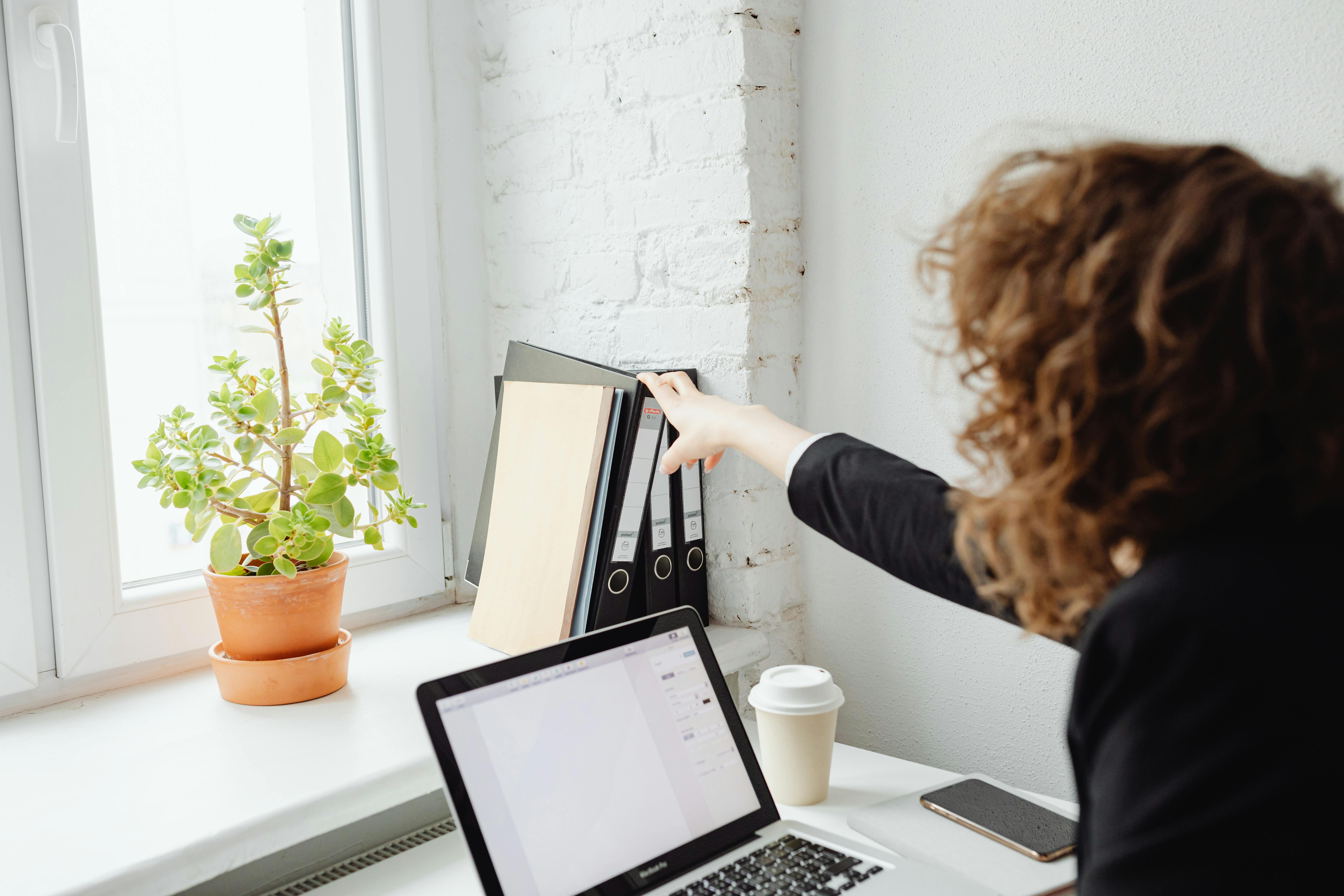 A woman reaches for folders in a light-filled modern office with a laptop.