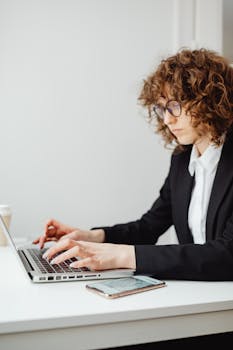 Focused young woman in business attire typing on a laptop at a desk indoors.