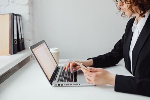 Free Professional woman in business attire using laptop and smartphone at a desk in an office setting. Stock Photo