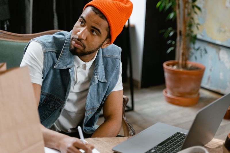 Young man in beanie focused on work with laptop and notebook in a modern boutique setting.