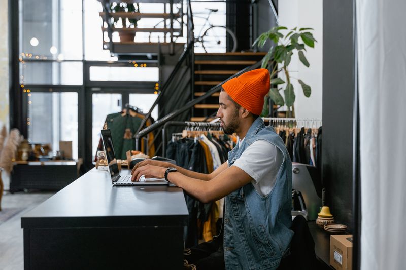 Shop owner working at a laptop behind the counter of a clothing store — photo by MART PRODUCTION on Pexels