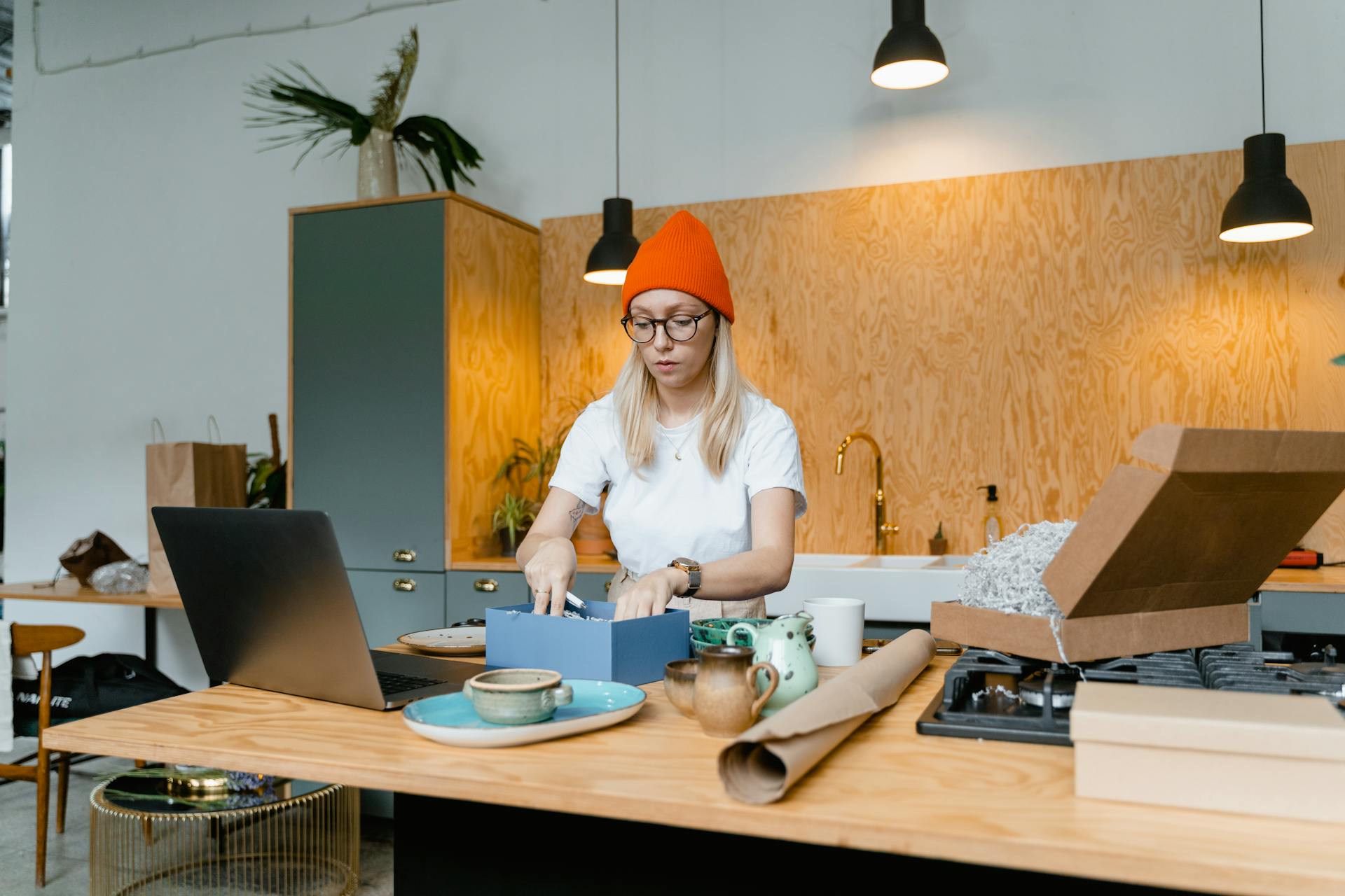 A woman in a kitchen packs a box while using a laptop, surrounded by kitchenware.