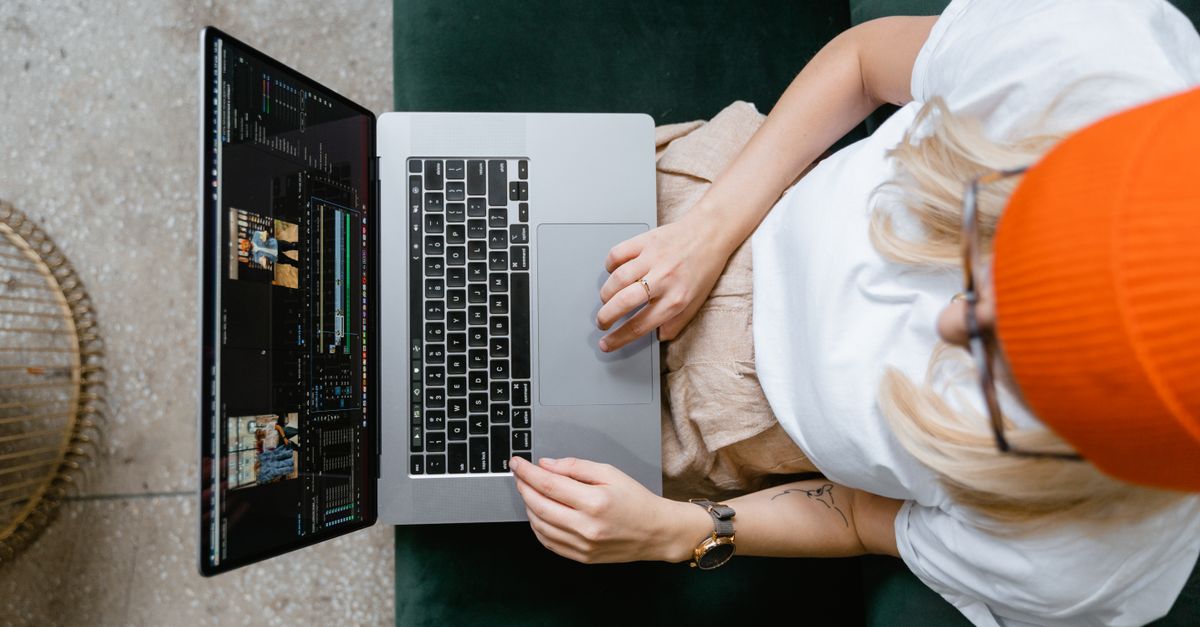 Top view of a woman editing video content on a laptop, wearing a casual outfit and orange beanie.