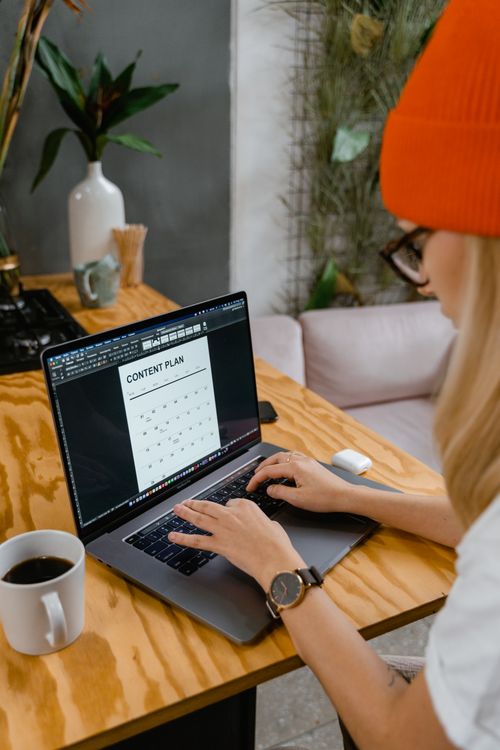 Free A woman wearing a knit hat types on a laptop at a wooden table with a cup of coffee indoors. Stock Photo