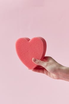 A hand holding a heart-shaped sponge with a soft pink background, symbolizing love and care.
