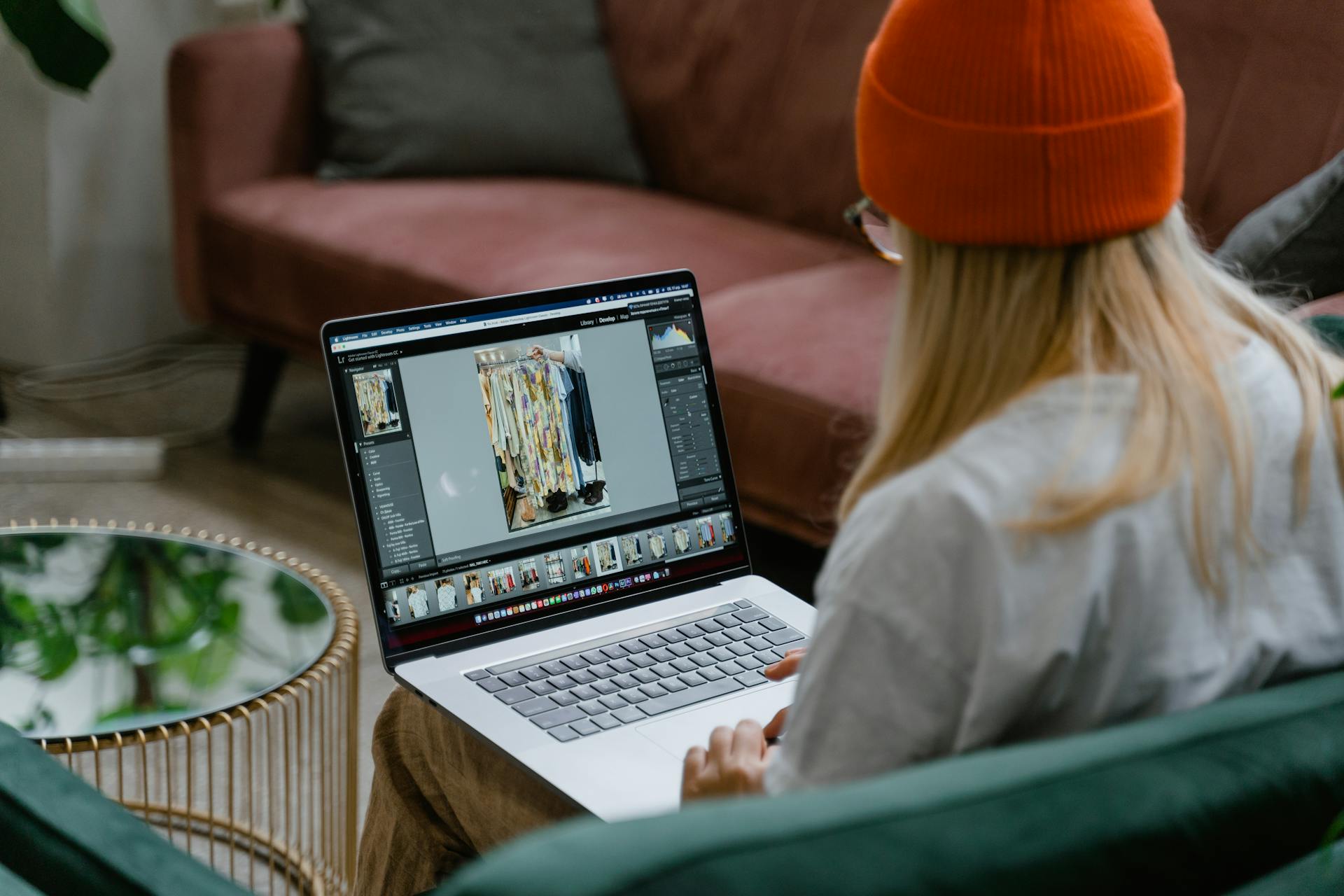 A woman wearing a beanie edits fashion photos on a laptop while seated on a cozy sofa.