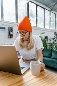 Young woman wearing orange beanie using laptop while holding a mug indoors, exuding a casual and focused vibe.