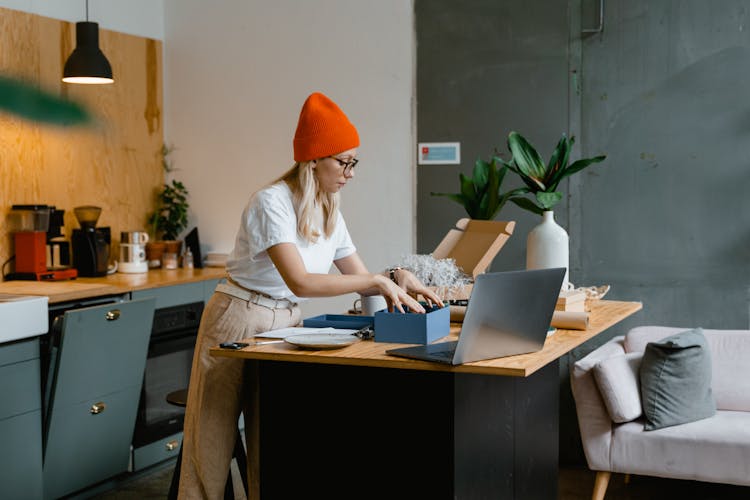 Young Woman Standing In The Kitchen With A Laptop And Unpacking An Order 