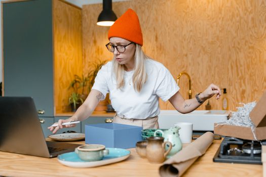 Focused woman in beanie working with laptop and ceramics in cozy home kitchen.