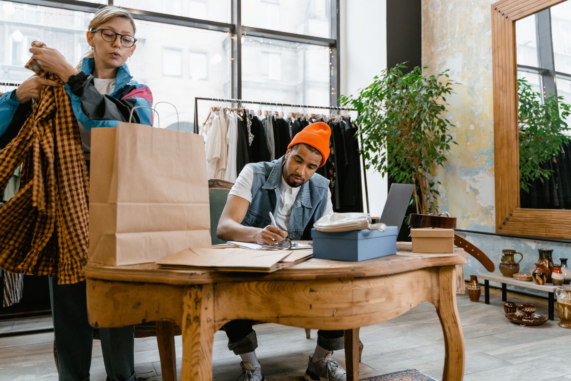 A Woman Putting the Clothes Inside the Paper Bag while Standing Beside a Man Working on His Desk