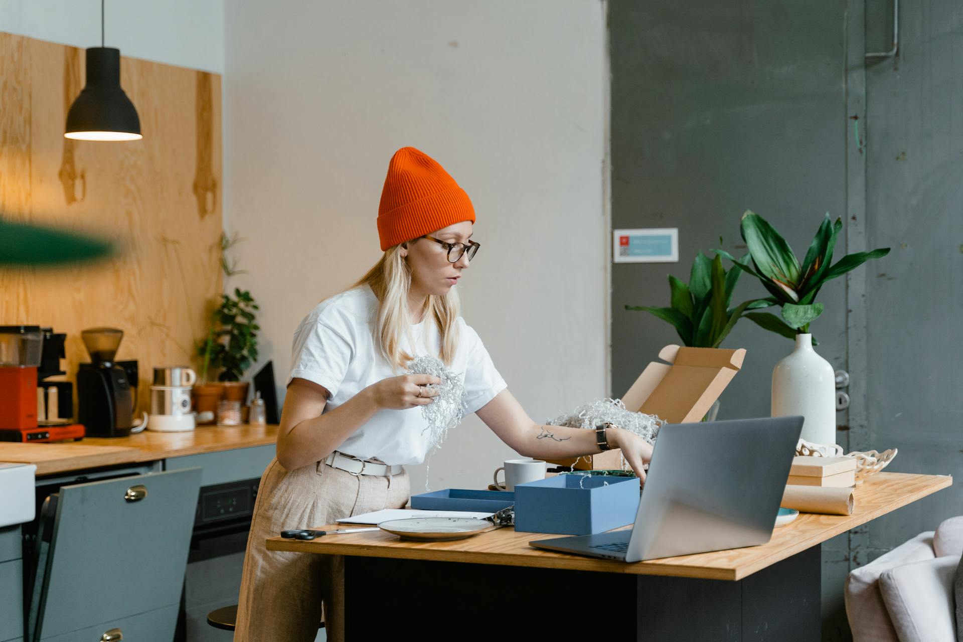 A woman wearing a beanie works from home packing an online order with a laptop and coffee nearby.