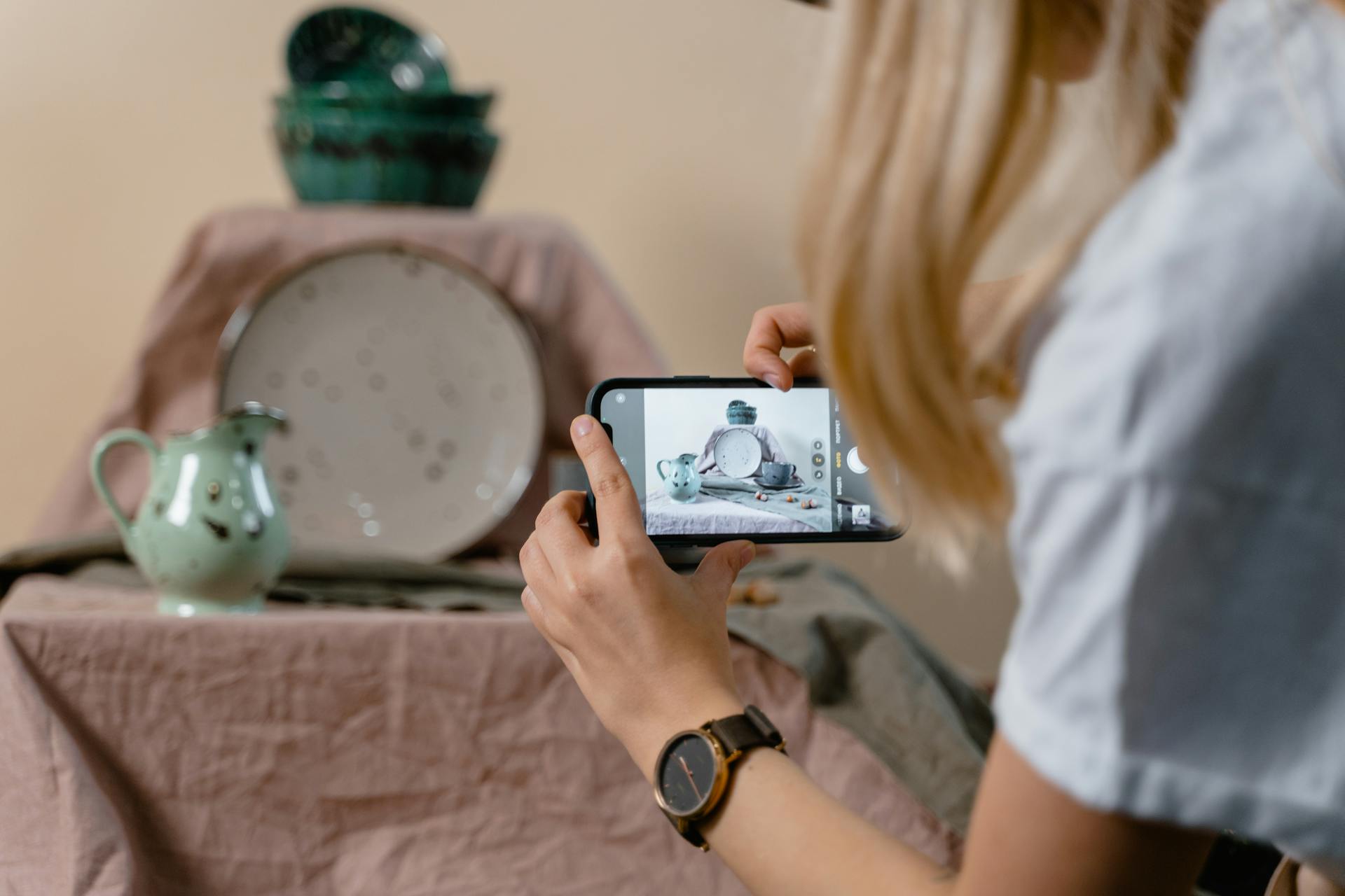 Person photographing elegant ceramic crockery setup with mobile phone for online selling.