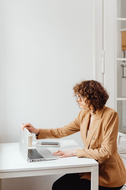 Free A woman in a brown blazer works on a laptop at an indoor office setting with a white desk. Stock Photo