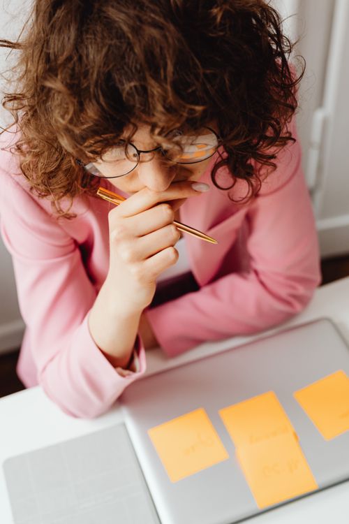 Free Woman in pink blazer pondering over laptop with notes, deep in thought at workspace. Stock Photo