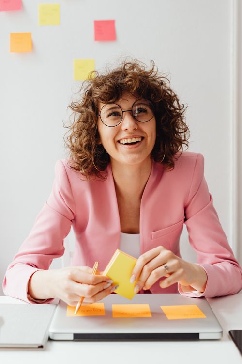 Free Cheerful woman in pink blazer with curly hair smiles while working at desk with sticky notes. Stock Photo