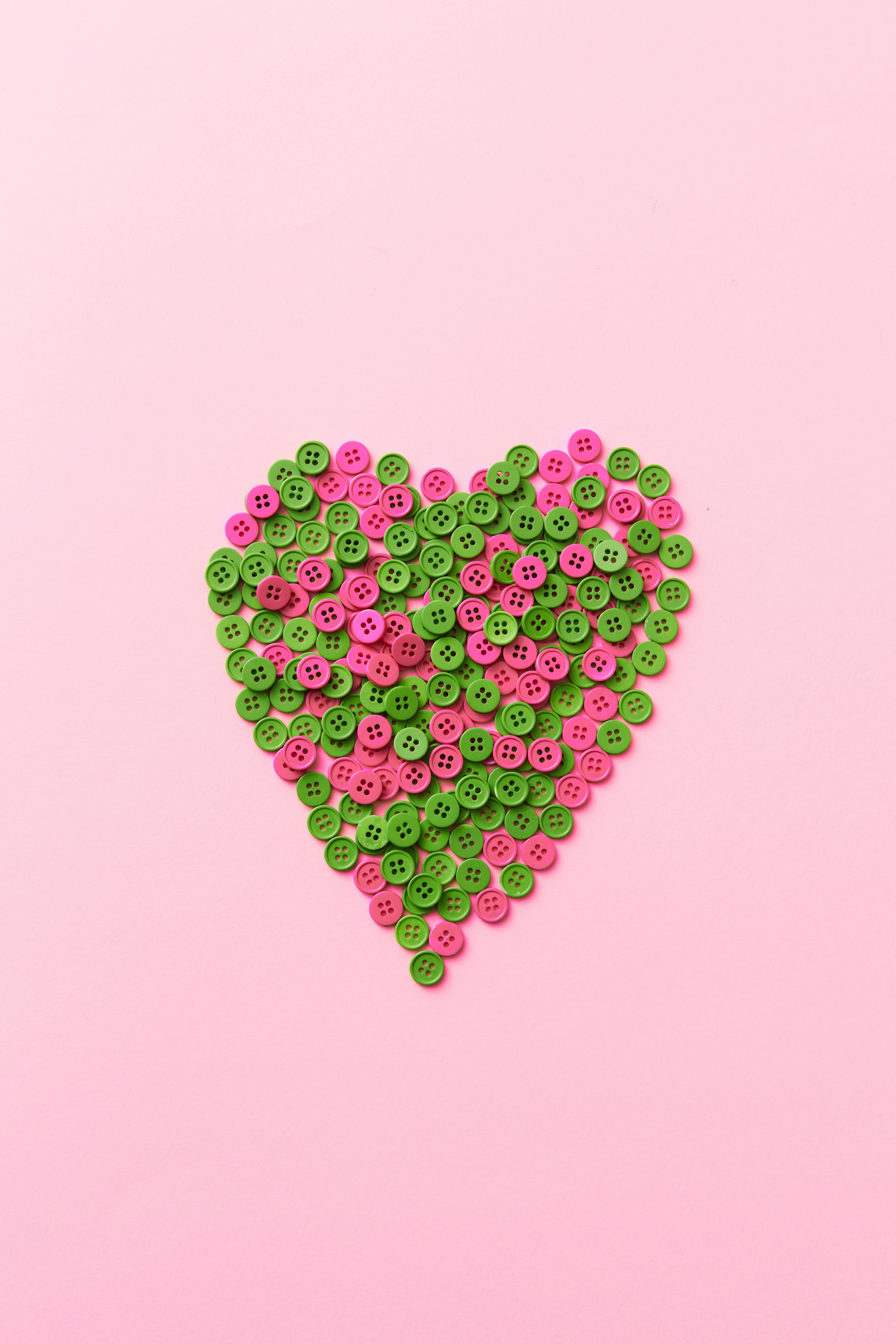 A heart shape created with green and pink buttons on a pastel pink background, radiating love and creativity.