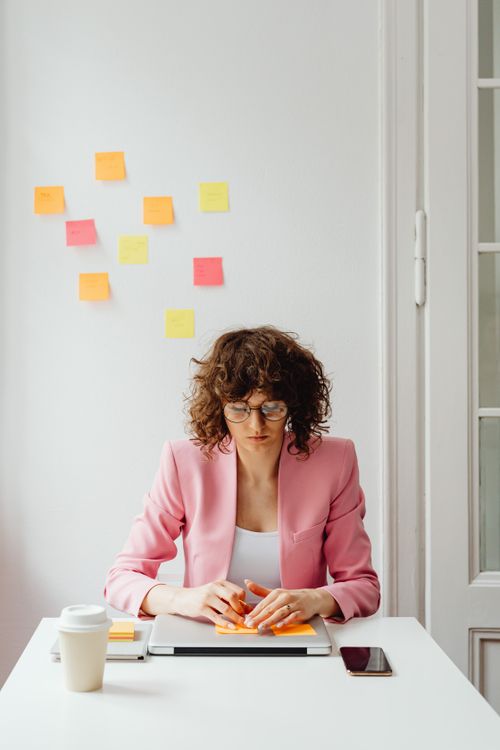 Free Businesswoman wearing a pink blazer focusing on work with sticky notes on wall in a modern office. Stock Photo
