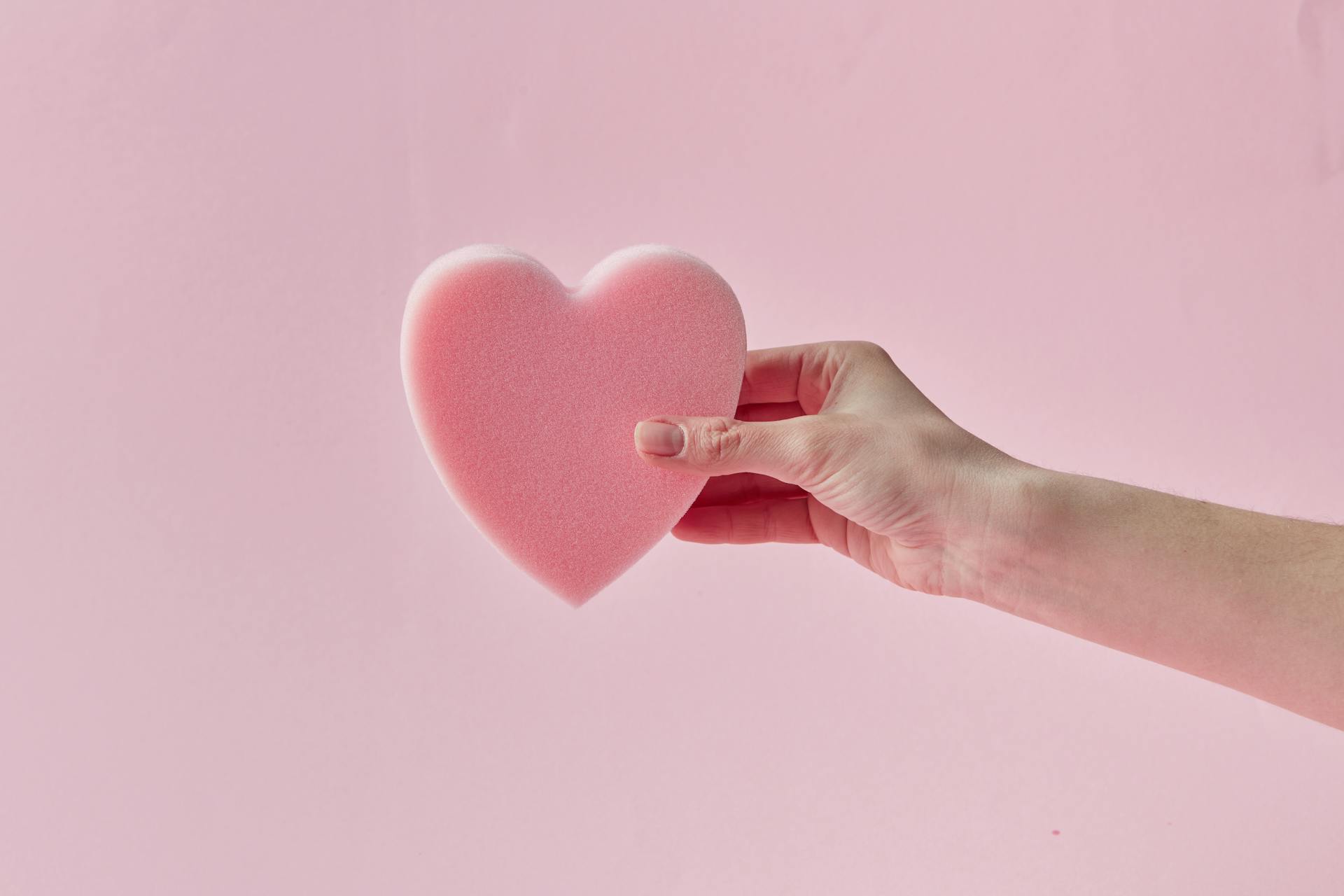 A heart-shaped foam held by a hand against a soft pink background, evoking love and tenderness.
