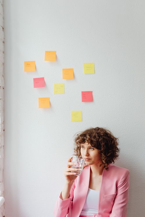 Free A woman in a pink blazer sits indoors, holding a glass of water, with sticky notes on the wall. Stock Photo