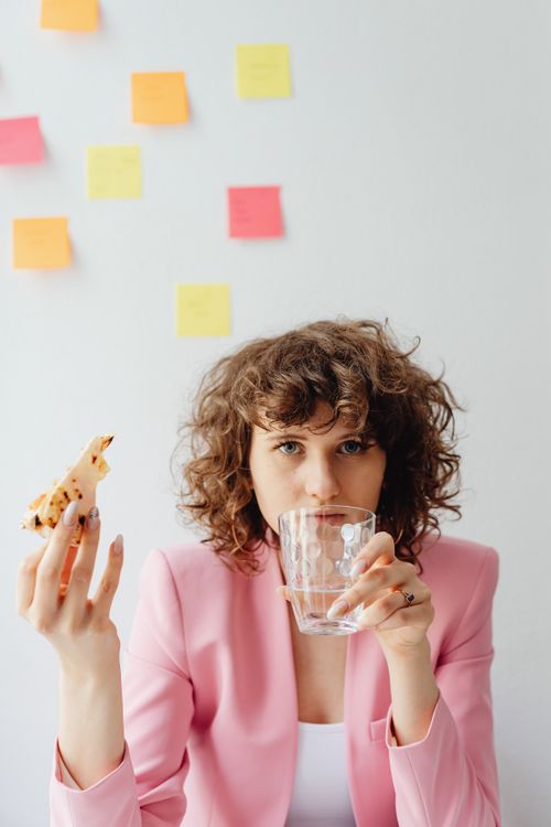 Free Caucasian woman with curly hair in pink blazer enjoying a break with pizza and water indoors. Stock Photo