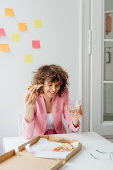 Businesswoman in pink blazer eating pizza during home office break. Relaxed and casual work environment.