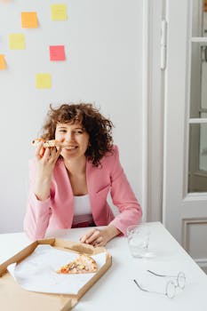 Smiling woman in pink blazer enjoys a pizza break at her desk surrounded by sticky notes.