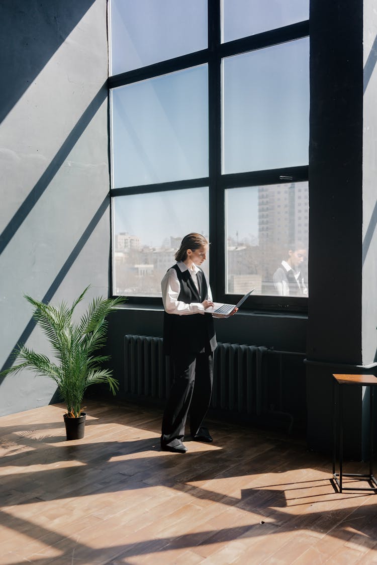 A Person Standing Near Glass Window While Typing On The Laptop She Is Holding