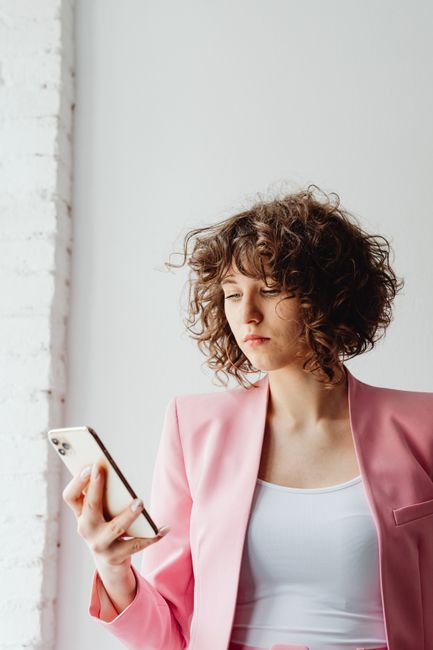 Curly-haired woman in a pink blazer using a smartphone indoors.