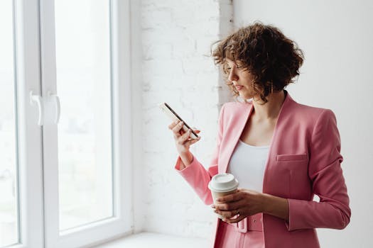 Young professional in pink suit checking smartphone while holding a coffee cup.