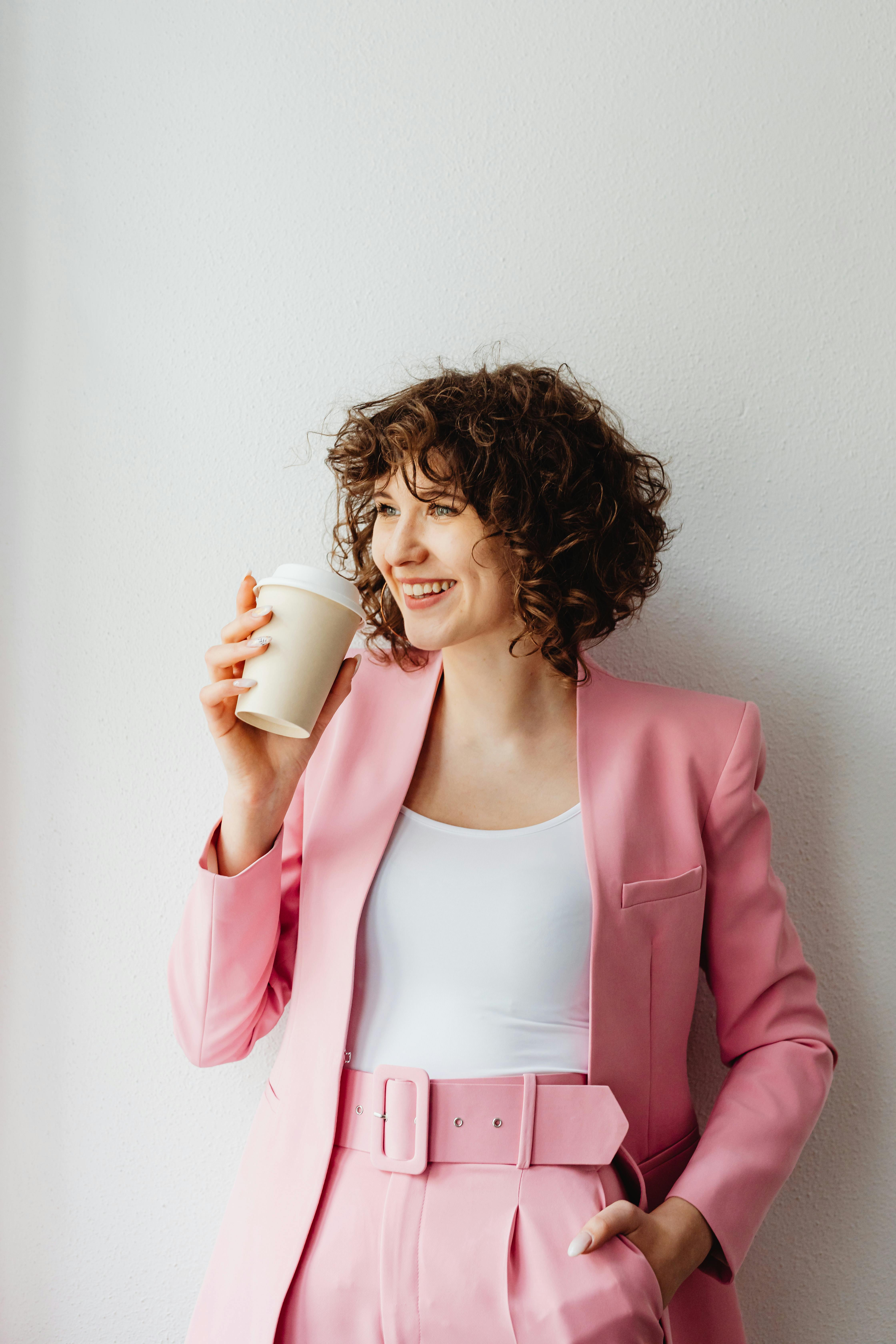 Confident woman in pink suit smiling with coffee cup indoors against white wall.