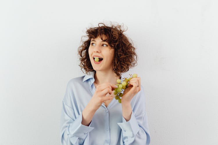 Curly-Haired Woman Eating Green Grapes On White Background