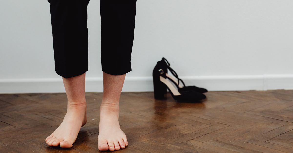 A person in black pants standing barefoot on wooden flooring next to black heels.