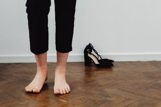 A person in black pants standing barefoot on wooden flooring next to black heels.