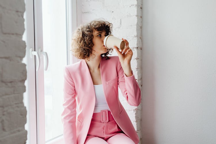 Woman In Her Pink Suit Drinking Coffee While Sitting By The Window