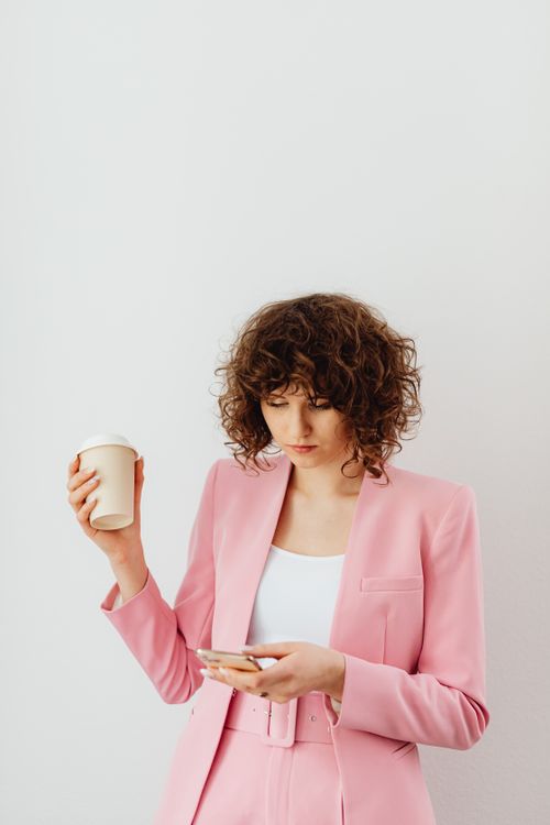 Free Confident woman in a pink blazer holding coffee and texting indoors. Stock Photo