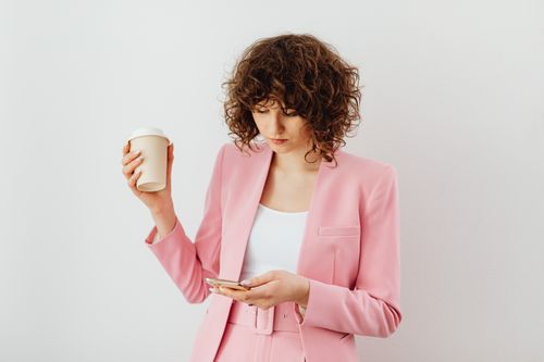 Free Stylish woman in pink suit holding coffee and using phone indoors. Stock Photo