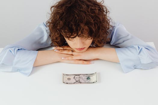 Caucasian woman with curly hair pondering over a five-dollar bill, expressing financial worry.