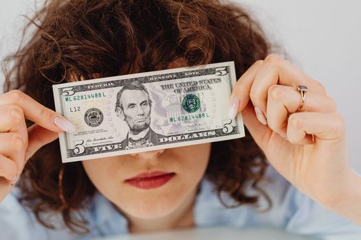 Woman with curly hair playfully covering eyes with a five dollar bill.