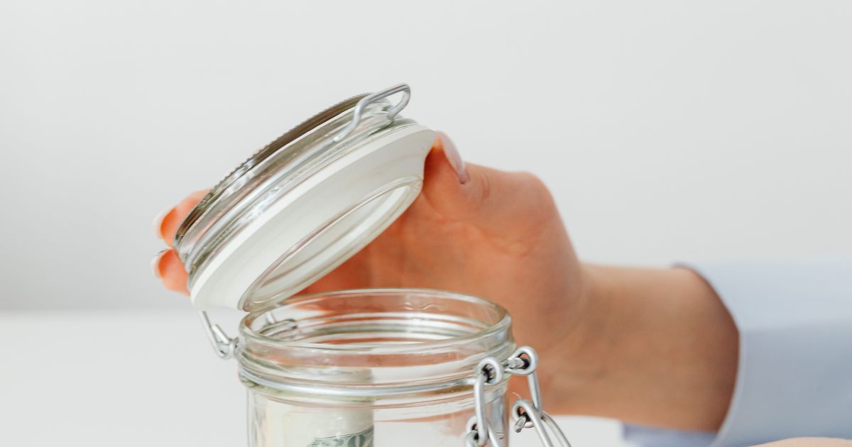 Manos depositando billetes en un frasco de ahorro sobre una mesa blanca, representando la aportación mensual automática al fondo de emergencia