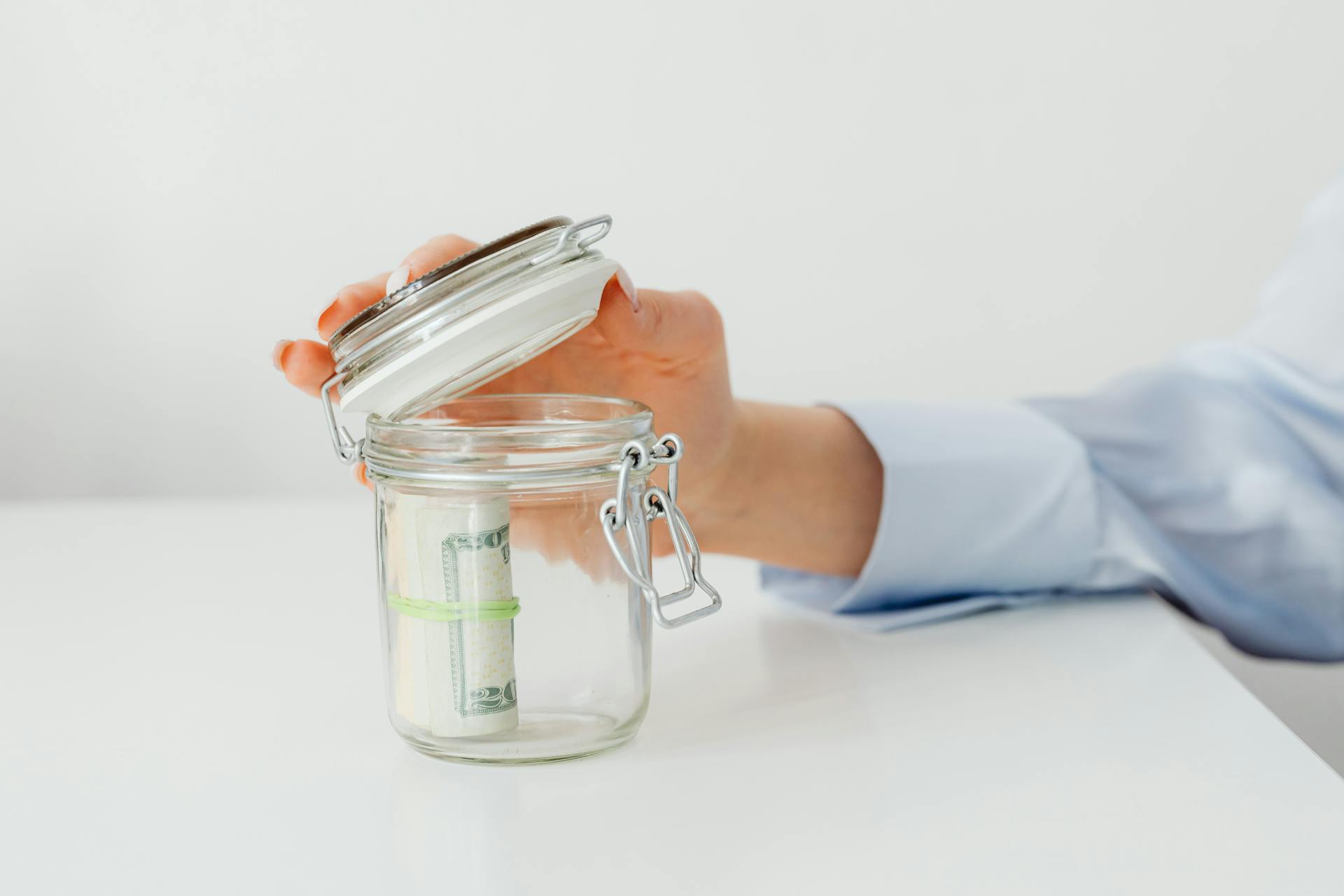 A hand places money in a glass jar on a white table, symbolizing savings.